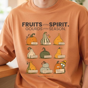 May include: An orange t-shirt featuring the text FRUITS of the SPIRIT. GOURDS of the SEASON. and nine illustrated gourds with faces, each holding a sign labeled with virtues like LOVE, JOY, PEACE, PATIENCE, KINDNESS, GOODNESS, FAITHFULNESS, GENTLENESS, and SELF-CONTROL.