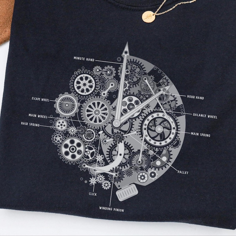 May include: Navy blue t-shirt with a detailed clock mechanism design in grey tones. The design includes labels like "Minute Hand," "Escape Wheel," and "Main Spring." A gold necklace is visible in the upper right corner.