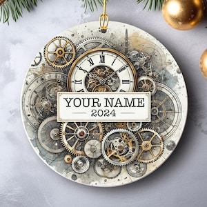 Könnte beinhalten: Ein weißer Keramik-Anhänger mit einem Steampunk-Design, das ein Zifferblatt und Zahnräder zeigt. Der Anhänger trägt den Text "YOUR NAME - 2024".