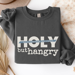 Könnte beinhalten: Dunkelgraues Sweatshirt mit den weißen Wörtern "HOLY" und "buthangry" und dem Satz "not by bread alone (but it helps)" in einem blauen Banner. Das Sweatshirt ist aus weichem Material.
