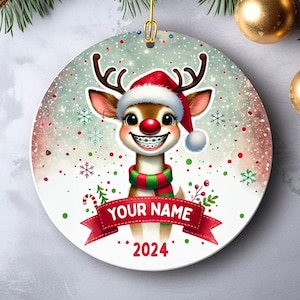 May include: A Christmas ornament featuring a cartoon reindeer wearing a red Santa hat and braces. The reindeer is smiling and has a green and red scarf. The ornament is decorated with snowflakes and red and green dots. The text "YOUR NAME" and "2024" are printed on the ornament.