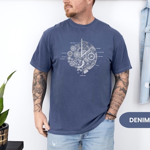 May include: A navy blue t-shirt with a white graphic of clock gears and mechanisms. The design is centered and includes labeled components. The shirt is made of a soft material. The word "DENIM" is in a blue oval.