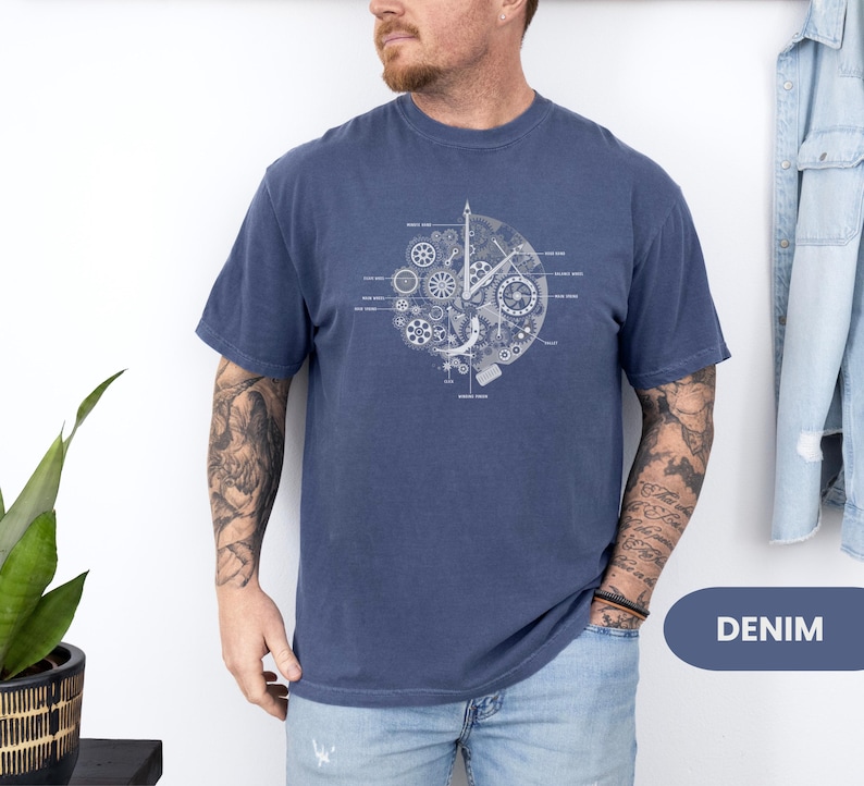 May include: A navy blue t-shirt with a white graphic of clock gears and mechanisms. The design is centered and includes labeled components. The shirt is made of a soft material. The word "DENIM" is in a blue oval.