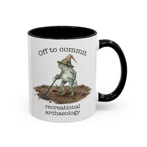 May include: White ceramic mug with a black interior and handle. The mug features a whimsical illustration of a frog with a metal detector, wearing a hat and headphones. Text on the mug reads "Off to commit recreational archaeology."