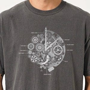 May include: A charcoal gray t-shirt featuring a white graphic print of a mechanical watch movement. The design includes detailed gears, springs, and labels such as minute hand, hour hand, balance wheel, main spring, pallet, click, and winding pinion.