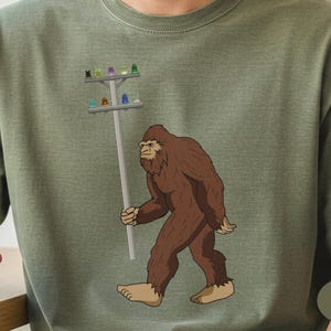 May include: An olive green t-shirt featuring a graphic illustration of a brown, furry Bigfoot creature walking while carrying a grey utility pole. The pole is topped with a crossbar holding several small, colourful glass insulators in various hues.