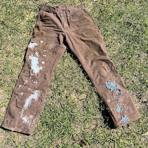 May include: Brown work pants with distressed patches of light blue fabric. The pants are laid out on a bed of green grass, showcasing a worn and rugged aesthetic. The pants have a classic five-pocket design.