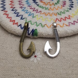 May include: Two metal fishing hook charms, one silver and one bronze, on a white background with a colourful woven pattern.