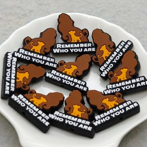 May include: A selection of lion-shaped charms in brown and yellow, each bearing the white text "REMEMBER WHO YOU ARE" on a black base. They are displayed on a white, shell-shaped dish.