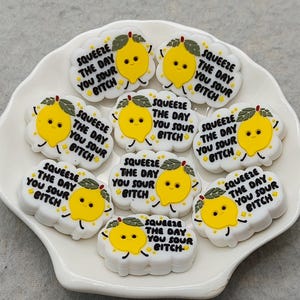 May include: A collection of white, cloud-shaped charms with a yellow lemon graphic and the text "SQUEEZE THE DAY YOU SOUR BITCH". The charms are arranged on a white, scalloped plate.