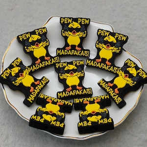 May include: A collection of black and yellow cartoon chick-shaped accessories with the words "PEW PEW" and "MADAFAKAS!" printed on them. The accessories are arranged on a white plate with a gold trim.