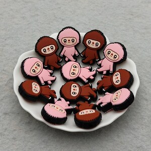 Puede incluir: Una colección de amuletos de perezosos de estilo caricatura en varias poses, dispuestos sobre un plato blanco con borde festoneado. Los perezosos tienen esquemas de color marrón y rosa con contornos negros. El plato está sobre una superficie gris.