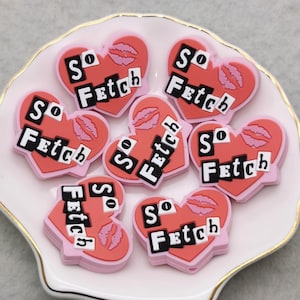 Puede incluir: Un conjunto de siete dijes en forma de corazón rosa con el texto "So Fetch" en letras negras. Cada dije tiene una impresión de labios rosa.