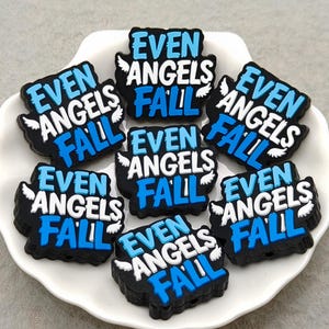 May include: A collection of black and blue shoe charms, each bearing the text "EVEN ANGELS FALL" in white and blue. The charms are displayed on a white plate. Each charm features small white wings.