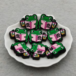 Op de afbeelding: Een verzameling groene kikkerbedels, elk met een roze boek en een drankje. De bedels zijn gerangschikt op een wit bord met geschulpte rand. De kikkers hebben zwarte contouren en staan tegen een lichtgrijze achtergrond.