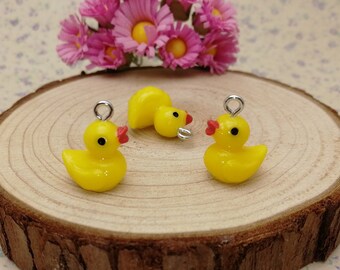 May include: Three small, yellow duck charms with red beaks and black eyes sit on a wooden slice. Each charm has a silver loop at the top. Pink flowers are in the background.