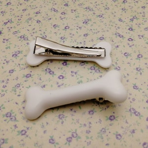 May include: Two white plastic bone-shaped hair clips. The clip on the left has a silver metal spring.