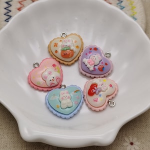 May include: Five heart-shaped charms with bunny and bear designs. The charms are in various pastel colors, including pink, blue, and purple. Each charm has a silver-colored loop at the top for attaching to a necklace or bracelet.
