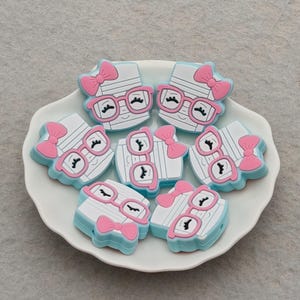 May include: A collection of white, blue, and pink decorative embellishments shaped like a face with glasses and a bow. The embellishments are arranged on a white, scalloped-edge plate against a gray background.