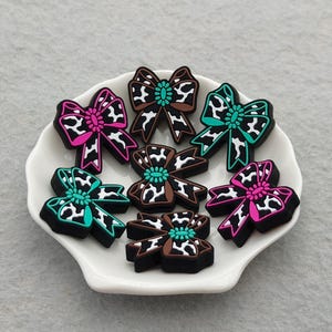 Op de afbeelding: Een verzameling van zes strikvormige bedels met een zwart-wit koeienprintpatroon. Elke bedel is voorzien van een effen gekleurd middenstuk en een bijpassende rand in felroze of levendig turquoise, gerangschikt op een wit schaaltje met schulprand.