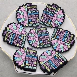 Op de afbeelding: Een set van zes kleurrijke, zonnebloemvormige bedels met de tekst "They whispered to her you can't withstand the storm she whispered back I am the storm", die op elke bedel wordt herhaald.