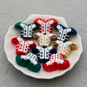 May include: A collection of miniature football jersey-shaped charms in various colors, including red, green, blue, and tan, with white accents. The charms are arranged in a white, shell-shaped dish.