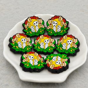 Op de afbeelding: Zeven kleurrijke schoenbedels met een Kodama-geestontwerp. Elke bedel heeft een gele achtergrond met een witte geestfiguur, omringd door groene bladeren en rode en gele bloemen. De bedels liggen op een wit, geschulpte bord.
