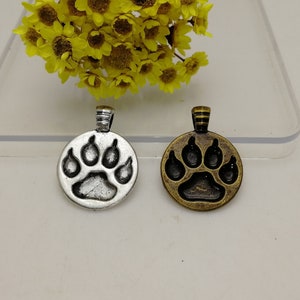 Puede incluir: Dos abalorios de plata y bronce en forma de huella de pata. Los abalorios son redondos y tienen un bucle en la parte superior para sujetarlos a un collar o pulsera.