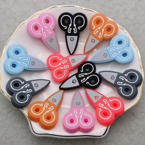 May include: A collection of 10 colourful silicone scissors charms in various colours, including pink, blue, orange, black, and grey. The charms are shaped like scissors with a decorative design and are perfect for adding a playful touch to jewellery, bags, or keychains.