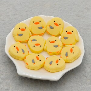 May include: A white, scalloped-edge dish filled with yellow chick-shaped and egg-shaped embellishments. The chicks have orange beaks and black eyes. The eggs are yellow with orange and black details. Perfect for crafting or Easter decor.
