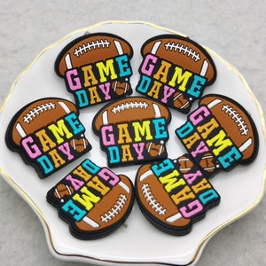 Może przedstawiać: Siedem brązowych i czarnych charmów w kształcie piłki nożnej z napisem "GAME DAY" w kolorowych literach. Charms znajdują się na białym talerzu ze złotą obwódką.