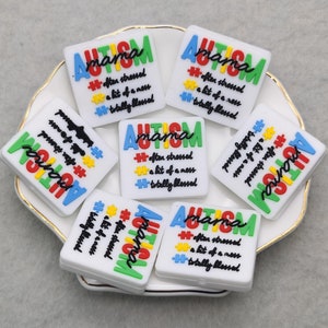 Könnte beinhalten: Ein Set aus acht weißen quadratischen Silikon-Anhängern mit dem Text "Autism mama" und dem Satz "after stressed, a bit of a mess, totally blessed" in bunten Buchstaben. Die Anhänger liegen auf einem weißen Teller mit goldenem Rand.