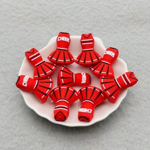 Puede incluir: Un conjunto de dijes con forma de falda de animadora en color rojo con rayas blancas y la palabra CHEER impresa en blanco. Estos pequeños artículos moldeados tienen un diseño acampanado y plisado con líneas negras y están en un plato festoneado.