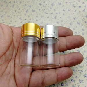 May include: Two small glass bottles with clear liquid inside. The bottle on the left has a gold lid with a silver band. The bottle on the right has a silver lid with a silver band.