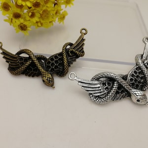 May include: Two metal snake charms with wings. One charm is bronze-coloured, the other is silver-coloured. Each charm features a snake coiled around a wing design, with a loop at the top for attaching to jewellery or other crafts.