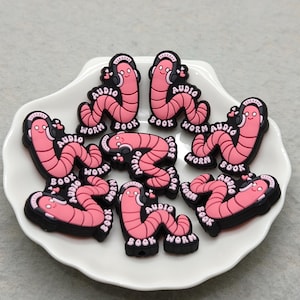 May include: A collection of pink worm-shaped charms with black outlines, each wearing headphones and the words "Audio Book Worm." The charms are arranged on a white, shell-shaped plate.