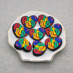 May include: A collection of heart-shaped charms with the word "PRIDE" in colorful letters. The charms are arranged on a white, scalloped dish with a gold rim. The letters are in rainbow colors.