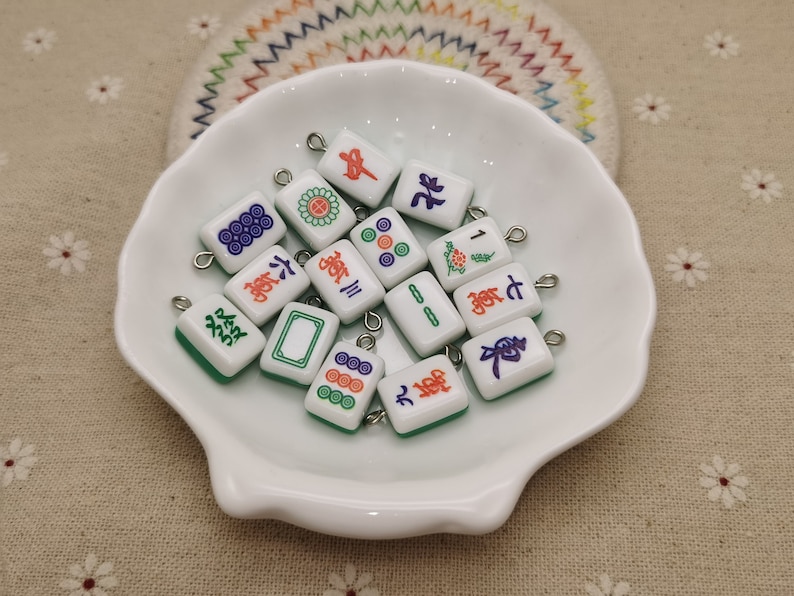 Peut inclure: Une collection de tuiles de mahjong en c&eacute;ramique blanche avec des caract&egrave;res et des symboles chinois verts, rouges et bleus. Les tuiles ont une boucle argent&eacute;e attach&eacute;e au sommet pour la suspension.