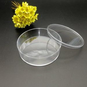 Puede incluir: Un recipiente redondo de plástico transparente con tapa. El recipiente está vacío y la tapa está quitada. El recipiente tiene un diámetro de unos 7,5 cm.