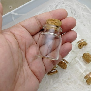 May include: A close-up of a small, clear glass bottle with a cork stopper. The bottle is being held in a hand, and there are several other similar bottles in the background.