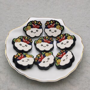 May include: Eight colorful cloud-shaped charms with the text "USA" and a red, white, and blue ribbon on top. The charms are on a white plate with a gold rim.