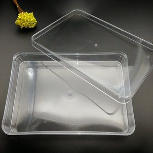 Puede incluir: Un recipiente de plástico transparente rectangular con una tapa a juego. La tapa está ligeramente entreabierta, revelando el interior del recipiente. El recipiente está diseñado para el almacenamiento y la organización. Un pequeño ramo de flores amarillas está en el fondo.