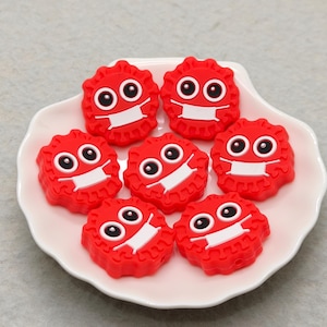 Puede incluir: Siete figuras rojas, tipo caricatura, con máscaras blancas y ojos negros, dispuestas sobre un plato blanco con bordes festoneados. Las figuras tienen una forma circular texturizada, que se asemeja a un virus o microbio.