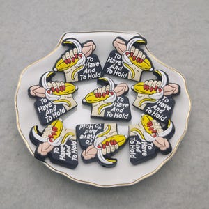 May include: A collection of banana-shaped charms with the text "To Have And To Hold" on a white seashell-shaped dish. The charms feature a yellow banana, a hand with red nails, and a black background.