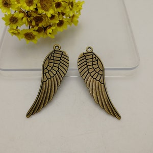 以下が含まれることがあります： 2つのブロンズ色の金属製の翼チャーム。各翼は詳細な羽のデザインで、ジュエリーやその他の工芸品に取り付けるための小さなループが上部にあります。翼はニュートラルな背景に表示されています。