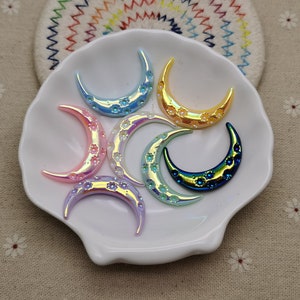 Puede incluir: Surtido de adornos de resina en forma de media luna iridiscente en varios colores, incluyendo azul, amarillo, rosa y morado. Los adornos tienen una superficie texturizada con pequeños puntos en relieve.