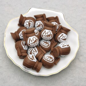 May include: A collection of brown silicone charms shaped like coffee pots with the words "Pot Head" printed in white on each charm.