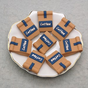 May include: Eight brown and blue coffee bag shaped charms with the word "Coffee" printed on each one. The charms are arranged on a white plate with a gold rim.