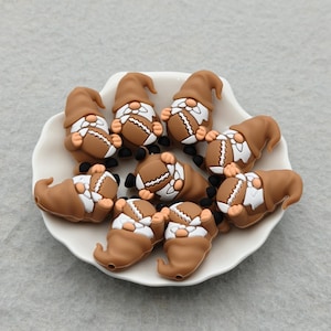 May include: A collection of brown gnome-shaped beads, each holding a football. The gnomes have brown hats and beards, with white and brown footballs. They are arranged on a white, scalloped-edge plate.