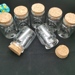 May include: Seven clear glass bottles with cork stoppers. The bottles are all the same size and shape, and they are arranged in a circle on a black surface.
